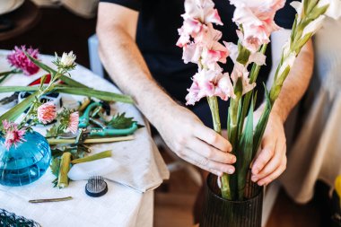Eller çalışma masasının üzerinde botanik süslemeler ve çiçeklerle kaplı büyük pembe ve beyaz gladiolus saplarını tutar. DIY çiçek tasarımı, yaratıcı beceri geliştirme, hobici çiçekçilik.