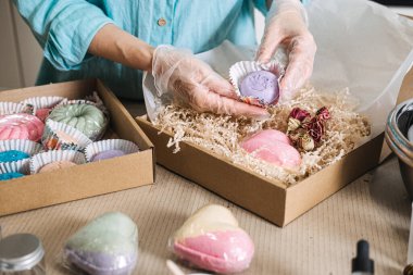Koruyucu eldiven giyen biri el yapımı mor banyo bombasını kağıt parçalarıyla dolu bir hediye kutusuna koyar. Sanatsal iş, ev temelli girişim, zanaat endüstrisi.