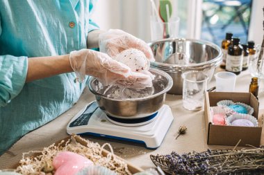 Elleri, metal kasenin üzerinde, ambalaj malzemeleriyle çevrili bir şekilde yuvarlak bir banyo bombası tutuyor. Banyo bombası yapımı, el yapımı kozmetik ürünler, sanatsal sabun, küçük parti güzellik, DIY banyo ürünleri.