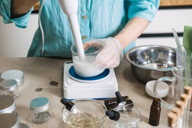 Dijital ölçekte beyaz sıvıyla doldurulmuş cam şişenin içine daldırma blenderi tutan kişi. Saf güzellikten rafine güzelliğe, organik kozmetik dönüşüme el yapımı cilt bakımı, botanik ürün geliştirme