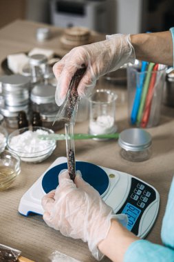 Dijital ölçekte bir test tüpünden diğerine koyu botanik malzemeler döken kişinin üst görüntüsü. Butik güzellik üretimi, mikro üretim, el yapımı bir markaya ölçeklendirme.