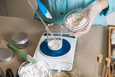 Eller, beyaz tozu dijital ölçekte cam şişeye aktarmak için mavi spatula kullanır. Laboratuvar stili estetik, kozmetik formülasyon süreci, dikkatli kimya, eczacı çalışma alanı