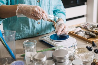 Eldivendeki eller, dijital ölçekte paketten cam test tüpüne koyu renkli botanik granüller döker. Yükseltilmiş Güzellik, Çöpten Hazineye, Tarım Atıkları Ajanları, Yükseltilmiş Botanikler