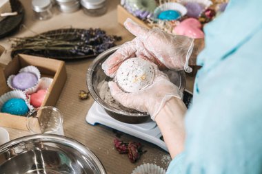 Eller beyaz tozu ve kurutulmuş botanikleri dijital ölçekte, bitmiş ürünlerle çevrili metal kasede karıştırıyor. El yapımı banyo bombaları, kozmetik kimya, sanatsal sabun yapımı.