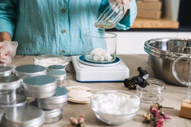 Eldivenin içindeki sıvı yağı dijital ölçekte beyaz katı tereyağı içeren kabın içine boşaltır. Kozmetik formülasyon süreci, yağ fazı emülsiyonu, krem doku değişimi, el yapımı nemlendirici.