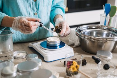 Person uses glass dropper to add oil to white cream in metal jar on digital scale. Lab-style beauty aesthetic, precise cosmetic formulation, diy apothecary lab, scientific skincare diy