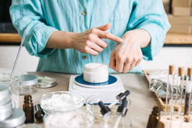 Person applies white cream to back of hand above jar on digital scale surrounded by skincare ingredients. tactile beauty DIY, silk-feel cream, sensory skincare testing, textural wellness ritual