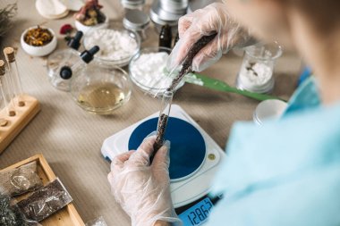 Dijital ölçekte bir test tüpünden diğerine koyu botanik malzemeler döken kişinin üst görüntüsü. Butik güzellik üretimi, mikro üretim, el yapımı bir markaya ölçeklendirme.