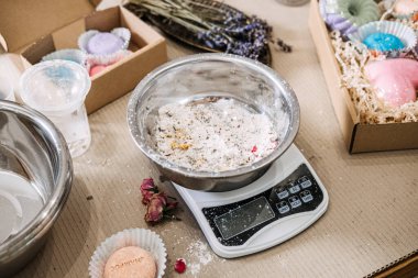 Metal bowl containing white powder and botanical inclusions sits on digital scale surrounded by packaging boxes. Cosmetic raw materials, ingredient study, clean formulation, skincare transparency