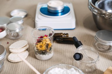 Kurumuş çiçekler ve toz içeren deney şişesi kozmetik kaplar ve ölçeklerin yanında iş yüzeyinde duruyor. Çiğ güzellik malzemeleri, cilt şeffaflığı, botanik özetleri, minimalist kozmetik malzemeler.