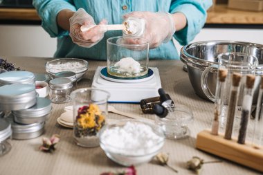 Hands in gloves scoop solid butter from jar into glass beaker on digital scale. raw beauty ingredients, skincare transparency trend, clean beauty formulation, botanical cosmetic actives