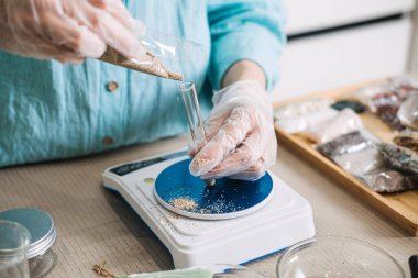 Eldivenli kişi bitkisel karışımı dijital ölçekte yerleştirilmiş cam test tüpüne döküyor. Modern botanik simya, laboratuardan banyoya ritüel, bilimsel doğa estetik, hassas bitki işçiliği