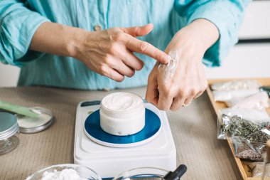 Kişi, dijital ölçekte beyaz kremi açık kavanozun üstüne, elinin tersiyle sürer. Zanaatkar parti testleri, zanaat estetiği, küçük ölçekli botanik boyası, el yapımı eko-lüks