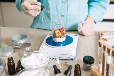 Person adds dried flowers to beaker on digital scale surrounded by cosmetic ingredients and jars. Zero waste beauty workshop, sustainable cosmetic production, eco-friendly DIY jars, small batch