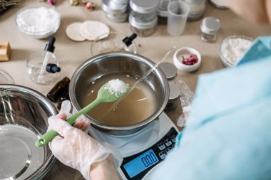 Person uses green spatula to hold white crystalline substance over metal bowl on digital scale. Hydration science aesthetic, pouring skincare liquid, water based cosmetic, refreshing beauty process