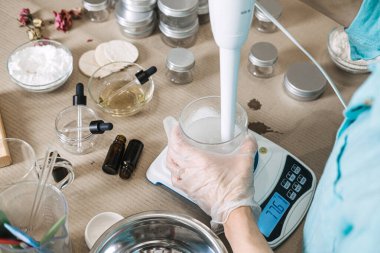 Dijital ölçekte beyaz sıvıyla doldurulmuş cam şişenin içine daldırma blenderi tutan kişi. Saf güzellikten rafine güzelliğe, organik kozmetik dönüşüme el yapımı cilt bakımı, botanik ürün geliştirme