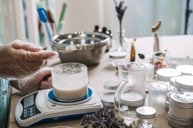 Person in gloves uses wooden spoon to add white solids into glass beaker on digital scale. Artisanal small batch beauty, luxury handmade skincare, exclusive apothecary drops, premium diy cosmetic