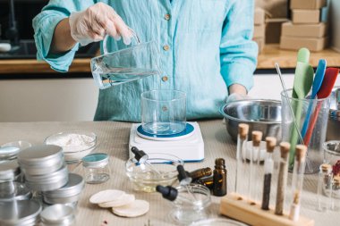 Person pours clear liquid from glass pitcher into glass beaker on digital scale. Cosmetic emulsion physics, oil and water mixing, skincare manufacturing science, professional cream base making