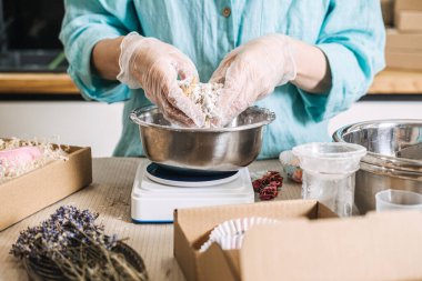 Person mixes botanical ingredients with hands in metal bowl on digital scale surrounded by packaging materials. Clean beauty process, transparent skincare brand, handmade cosmetic workshop.