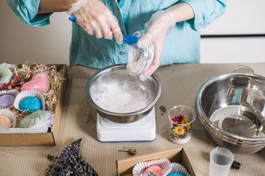 Person pours white powder into stainless steel bowl on digital scale surrounded by colorful finished products. Handmade bath bombs, personalized self-care gifts, bath salt mixology