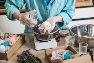 Hands hold large white bath bomb over metal bowl on digital scale surrounded by gift boxes. Small business scale, bulk bath bombs, production day vlog, handmade workshop