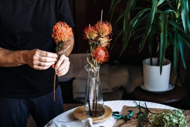 Masadaki cam vazoda parlak turuncu iğneli protea çiçeklerini düzenler. Koyu renk akademi çiçekleri, Cottagecore çiçek tasarımı, karamsar romantik buketler, klasik botanik estetiği