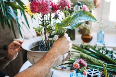 Adam pembe ve mor asterileri tel örgü kullanarak klasik beyaz vazoda düzenliyor. Dopamin dekoru çiçekleri, parlak çiçek enerjisi, moral yükseltici buketler, renkli çiçek terapisi..