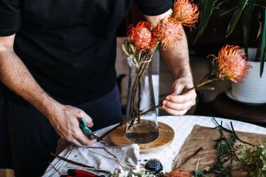Masadaki cam vazoda buketi düzenlerken portakal iğneli protea çiçeğini tutan kişi. Sanat terapisi, dokunsal farkındalık, duyusal sağlık, terapi amaçlı bahçıvanlık..