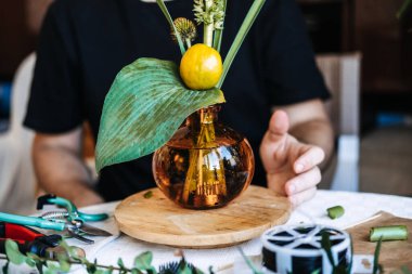 Çiçekçi, kehribar cam vazo taşıyan ahşap kaideyi botanik düzeniyle çeviriyor. Yavaş çiçek akımı, yerel çiçek yetiştiriciliği, doğaya bağlı yaşam tarzı.