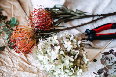 Çiçek ve çiçekçi aletleri kırışık kağıt ve açık kumaş üzerinde dinlenir. melez çiçek tasarımı, karışık dokular, kurumuş ve taze çiçekler, botanik kontrastı, modern çiçekçilik, çiçek koruma.