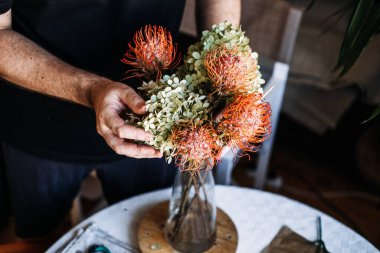 Eller canlı turuncu proteyi ve yeşil ortancayı masanın üstündeki cam vazoda düzenler. Doku Melezleri Kurumuş ve Taze Çiçekler, Canlı harmanlanmış taze bitkiler ve korunmuş elementler.