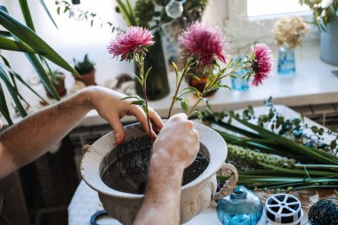 Eller pembe çiçekleri düzenlemek için tel örgüyü taş vazoya yerleştirir. Sürdürülebilir çiçek tasarımı, köpüksüz teknikler, yeniden kullanılabilir çiçek araçları