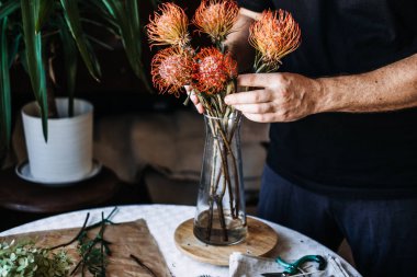Eller portakallı iğnelik protea çiçeklerini masanın üzerinde cam vazoya dönüştürür. el sanatları, çiçekçilik, lüks ev aksanları, el yapımı lüks, özel botanik sanatı.