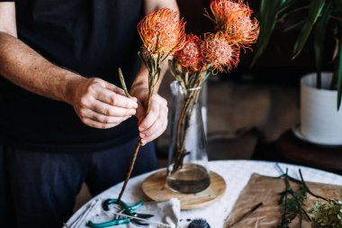 Elleri cam vazoya yerleştirmeden önce turuncu iğnelik protezin gövdesini budarlar. Zanaatkar çiçekçiler, lüks ev aksanları, üst düzey çiçekçiler, eşsiz iç mimari..