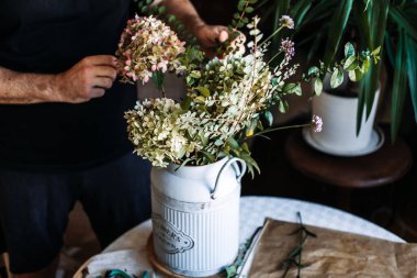 Eller, beyaz metal süt kutusunda ortanca ve yapraklı dalları düzenler. DIY çiçek aranjmanı, ev çiçekçiliği hobisi, yaratıcı eğlence, pratik öğrenme, el sanatları dersleri.