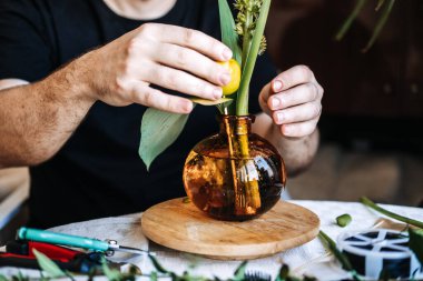 Eller, ahşap zemindeki kehribar cam vazodaki botanik elementleri düzenler. Çiçekçi, modern erkek hobileri, zanaatkar kimliği olan erkekler.