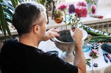 Adam, masadaki taş vazonun içindeki tel örgüye pembe paskalya sapları yerleştiriyor. Erkek bahçıvan hobisi, evde yetişen çiçekler erkekler, çiçekçi erkekler.