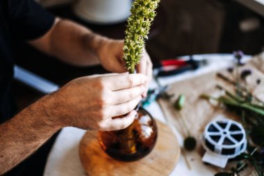 Eller masaya uzun yeşil çiçek sapını cam vazoya yerleştirir. Japon çiçek aranjmanı, minimalist estetik, meditasyon sanatı, zen yaşam tarzı, hassas çiçekçilik, kültürel miras.