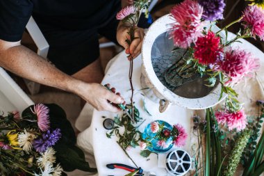 Eller, çiçek sapını kesmek için çiçek makası kullanır. Beyaz masa üzerinde çeşitli çiçekler ve vazolarla. dijital detoks yeşillik, kortizol indirme bitkileri, doğa terapisi kapalı alan.