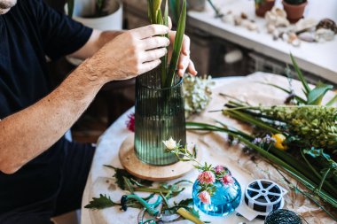 Çiçekçilerin elleri uzun yeşil sapları cam vazolara yerleştirerek masada aletler ve çiçek malzemeleriyle. Bağımsız çiçekçi, yerel işletme sahibi, küçük işletme girişimi, zanaat endüstrisi, çiçekçi dükkanı