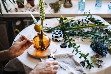 Eller sarı meyveyi kehribar cam vazonun yanında masanın üzerinde çiçek aranjmanıyla tutuyor. Çağdaş çiçek tasarımı, botanik sanatı, eşsiz bitki düzenlemeleri, sanatsal çiçekçilik.