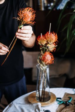 Masadaki cam vazoda parlak turuncu iğneli protea çiçeklerini düzenler. Koyu renk akademi çiçekleri, Cottagecore çiçek tasarımı, karamsar romantik buketler, klasik botanik estetiği