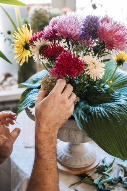 İnsanın elleri, canlı paskalya çiçekleri ve dekoratif vazoda büyük hosta yaprakları düzenler. Biyofili hipotezi, zihinsel iyileştirme, doğa destekli terapi, erkek psikolojik dayanıklılığı.