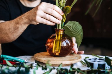 Eller, ahşap zemindeki kehribar cam vazodaki botanik elementleri düzenler. Çiçek tasarım kursu, yaratıcılık, mesleki beceriler ve zanaat eğitimi için sürekli eğitim