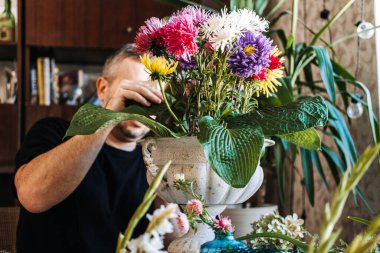 Adam renkli paskalya çiçekleri ve beyaz vazoda büyük hosta yaprakları düzenler. Odaklanma temelli terapi, erkekler için akıl sağlığı, hobi ustalığı, bilişsel refah.