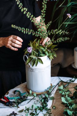 Eller, beyaz flüt sürahisinin içinde yapraklı saplar ve küçük mor çiçekler düzenler. Çiftlik çiçekleri şık, kır çiçekçisi, süt sürahisi, kır tarzı ev, pastoral estetik.