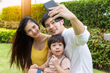 Genç aile, mutlu ve mutlu anıları korumak için telefon kullanarak fotoğraf çekiyor ve açık havada gülümsüyor. Baba, selfie çekmek için akıllı telefon kullanıyor ya da karısına ve küçük çocuğuna fotoğraf çekiyor. Mutlu aile kavramı.