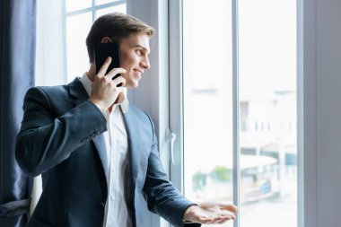 İş adamı, iş yerindeki pencerenin yanında duruyor. Takım elbiseli yakışıklı iş adamı, gülümseyen yüzüyle akıllı telefonla konuşuyor. Finansal İletişim konsepti kopyalama alanı.