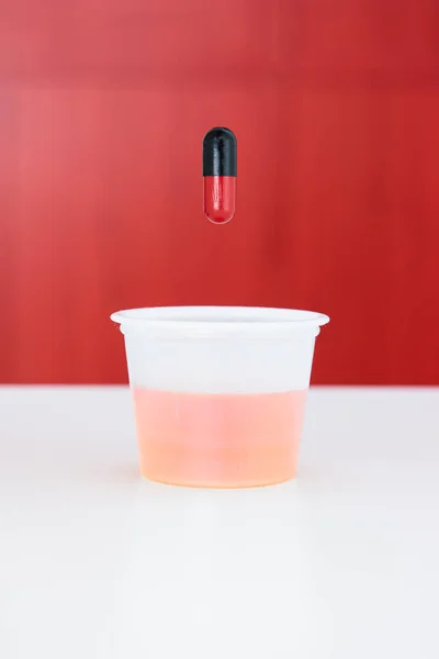 小杯透明塑料药杯 红色和黑色药物胶囊在其上方浅水区深度 — 图库照片