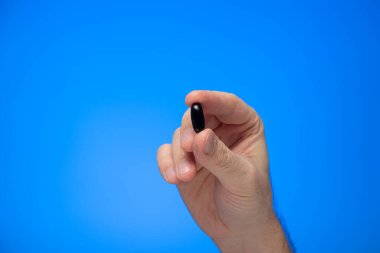 Kafkas erkek eli tarafından parmaklarının arasında tutulan siyah kapsül tıbbi hapı. Mavi arka plan stüdyo çekimlerinde izole edilmiş..